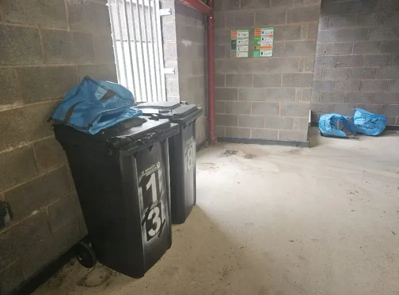 Two black wheelie bins and blue recycling bags in a concrete utility area, ideal for garage clearance or rubbish removal.